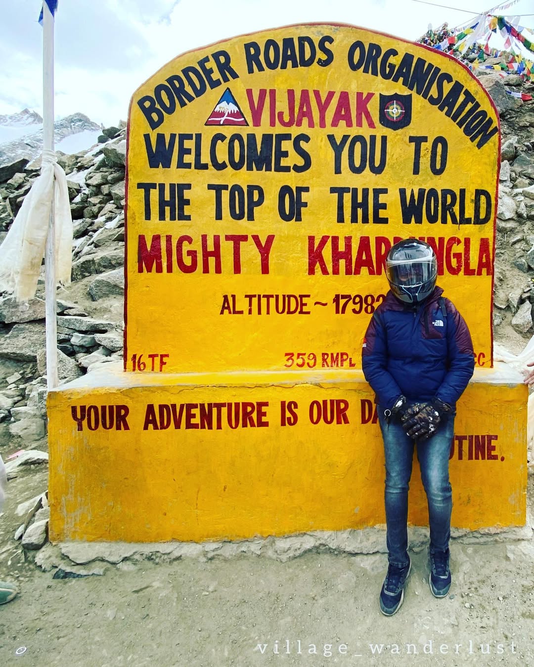 Thenmozhi wearing a blue jacket and motorcycle helmet beside the yellow Khardung La pass sign.