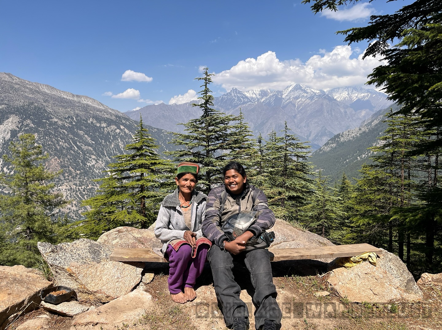 Thenmozhi seated beside a local woman, with pine trees and snowy mountains in Kinnaur.