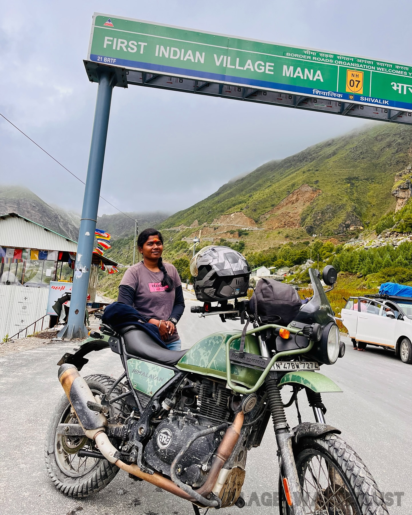 Thenmozhi and her green Royal Enfield Himalayan beneath the Mana village sign.