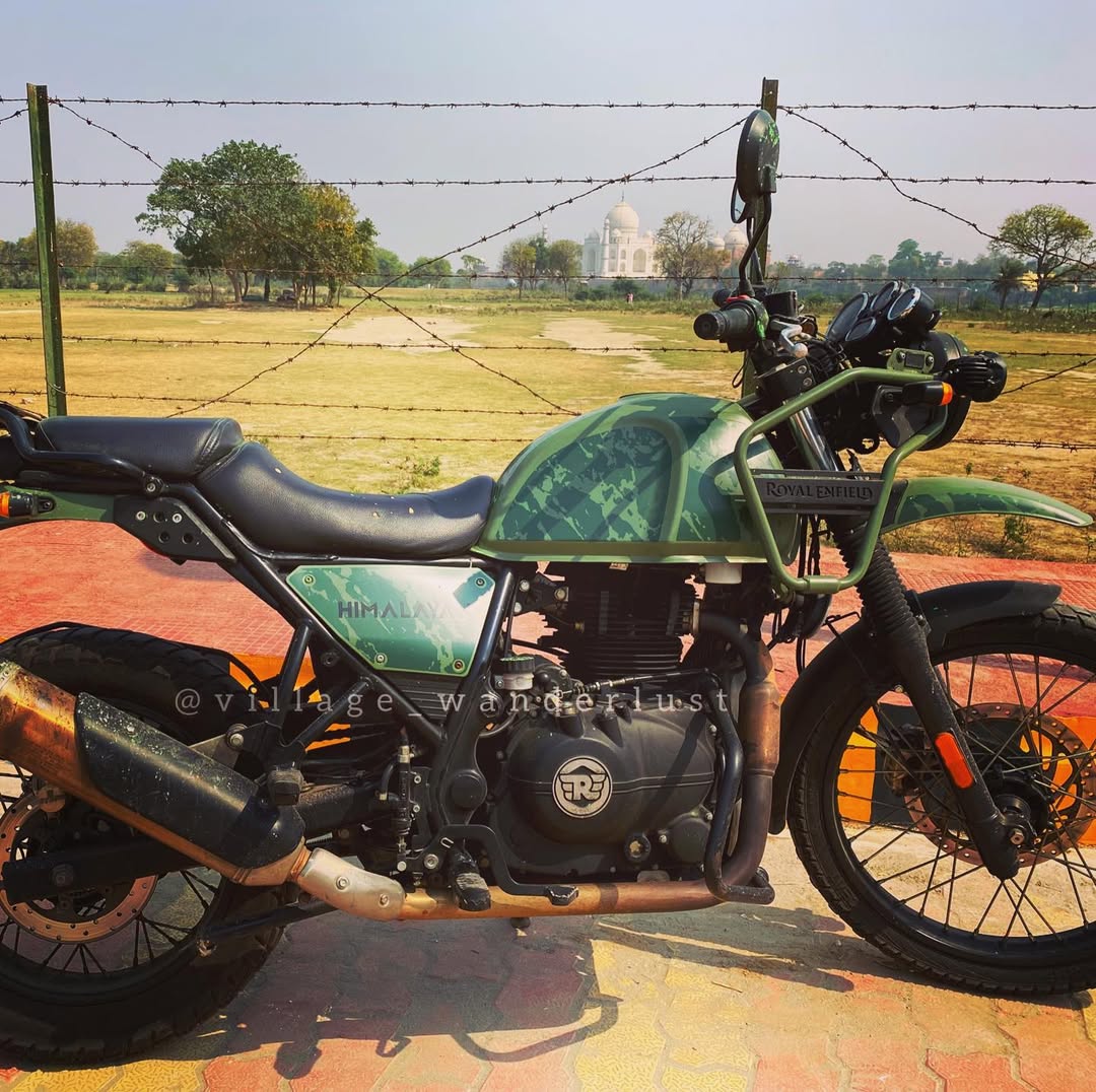 A green Royal Enfield Himalayan with the Taj Mahal visible beyond a fence in Agra.